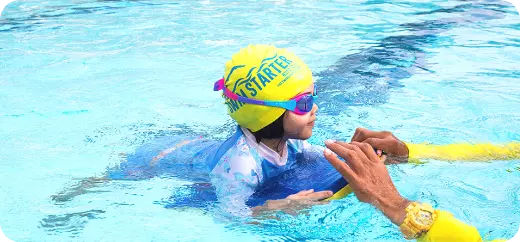 Child learning to swim with a yellow swimming board in the deep pool under the guidance of a Singapore swimming coach